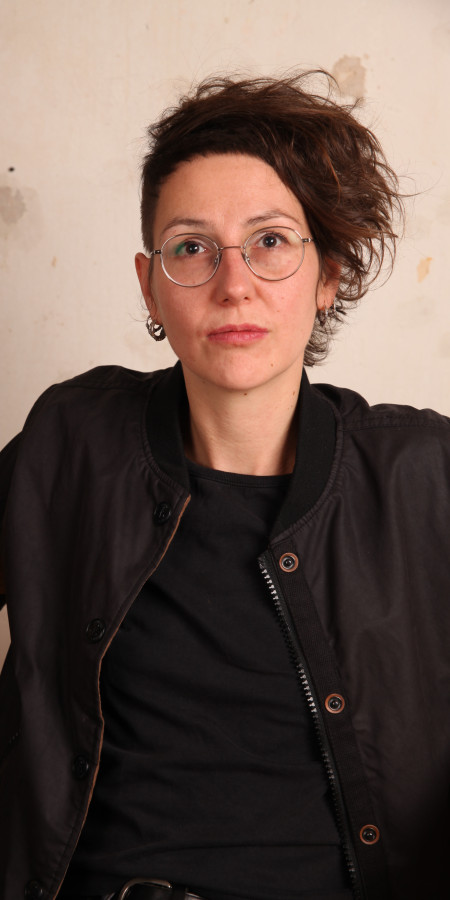 Das Bild zeigt ein Poträt von Jule Govrin. Eine weiblich gelesene Person mit asymetrischer Frisur und Brille. Sie trägt ein schwarzes Oberteil.