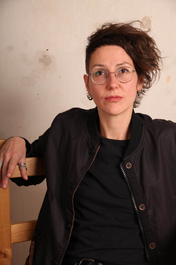 Das Bild zeigt ein Poträt von Jule Govrin. Eine weiblich gelesene Person mit asymetrischer Frisur und Brille. Sie trägt ein schwarzes Oberteil.