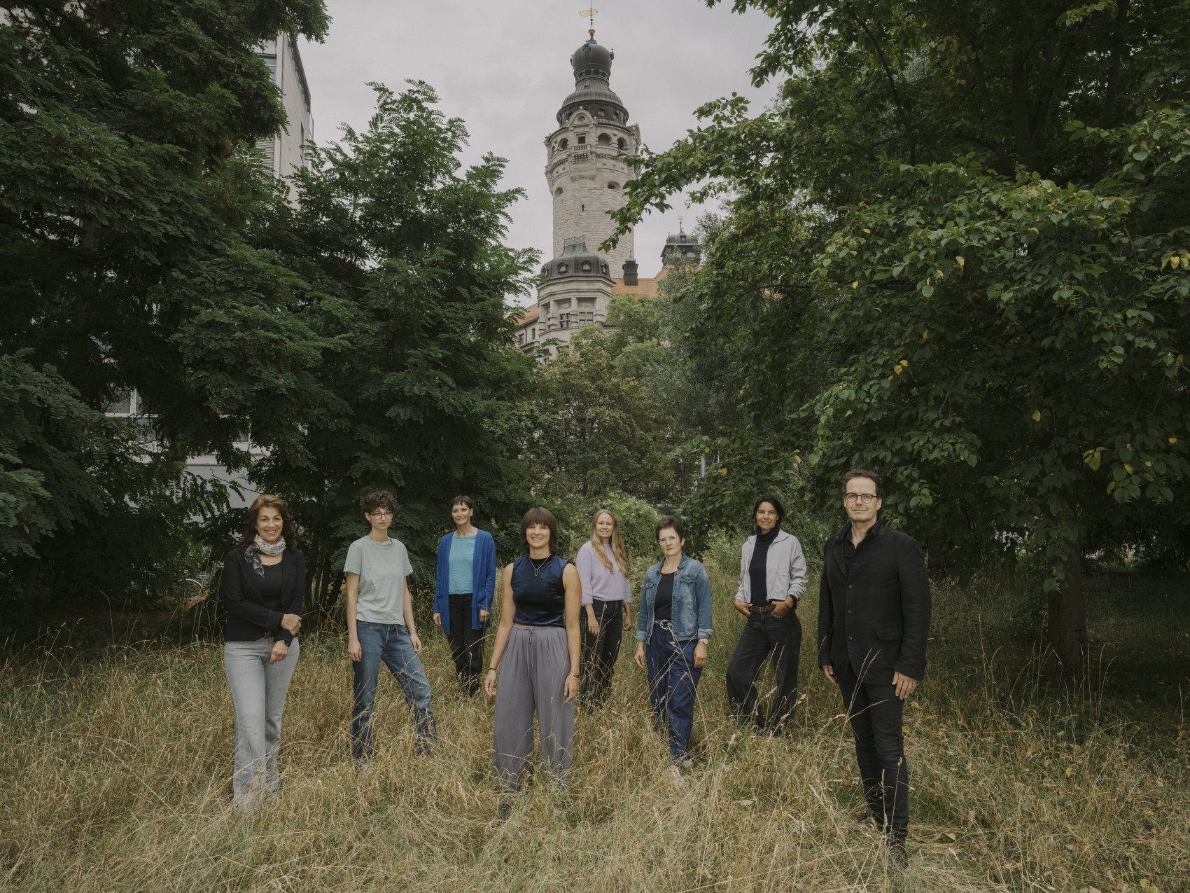 zu sehen ist das Team der euro-scene Leipzig 2026, 8 Personen auf einer Wiese mit dem Turm des Neuen Rathaus Leipzigs im Hintergrund