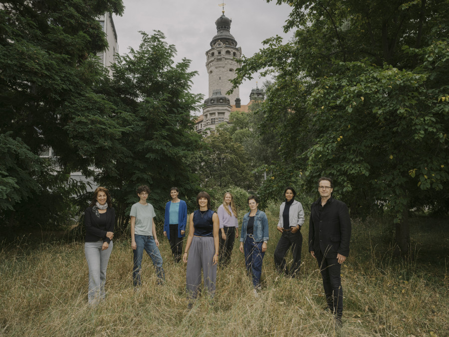 zu sehen ist das Team der euro-scene Leipzig 2026, 8 Personen auf einer Wiese mit dem Turm des Neuen Rathaus Leipzigs im Hintergrund