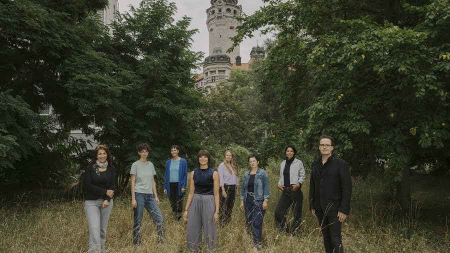 zu sehen ist das Team der euro-scene Leipzig 2026, 8 Personen auf einer Wiese mit dem Turm des Neuen Rathaus Leipzigs im Hintergrund