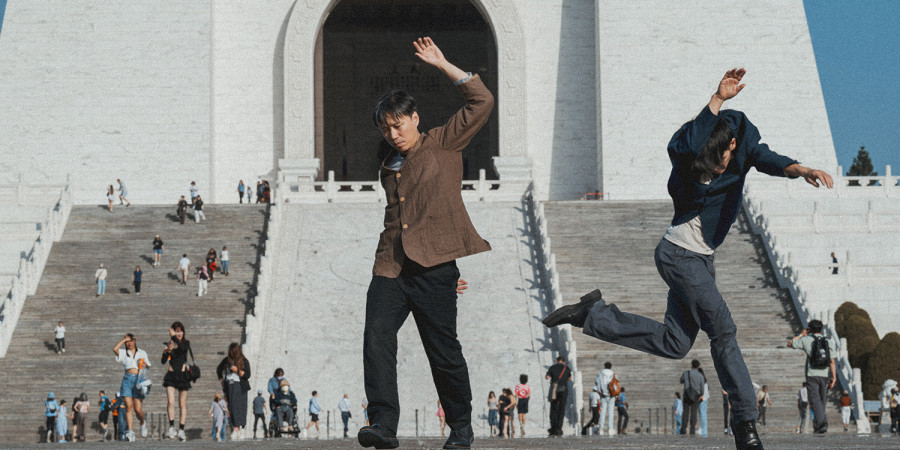 Das Bild zeigt die beiden Tänzer des Duett vor einem weißen Gebäude zu dem Treppen hinaufführen. Auf dem Platz sind im Hintergrund einige Menschen zu sehen. Beide Tänzer sind in einem niedrigen Sprung zu sehen.