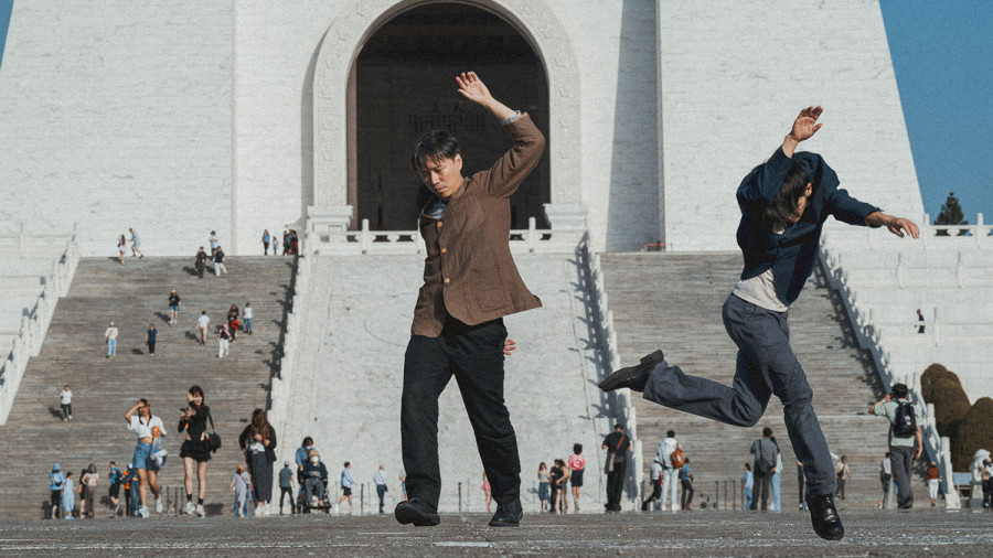 Das Bild zeigt die beiden Tänzer des Duett vor einem weißen Gebäude zu dem Treppen hinaufführen. Auf dem Platz sind im Hintergrund einige Menschen zu sehen. Beide Tänzer sind in einem niedrigen Sprung zu sehen.