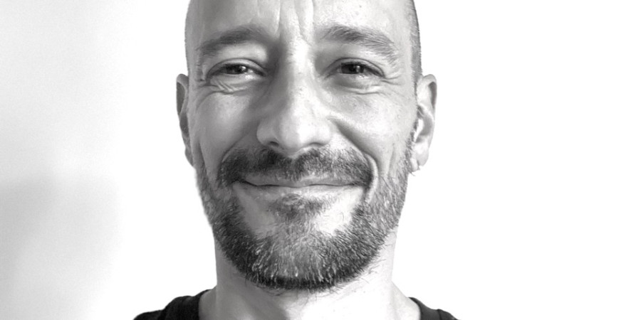 Das Bild zeigt ein Porträt des CHoreografen Manuel Roque. Ein weiße männliche gelesene Person ohne Haare mit leichtem Bart und breitem, freundlichen Lächeln.