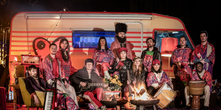 Das Bild zeigt den Caravan of LUV - insgesamt 12 Personen, männlich und weiblich gelesen vor einem Wohnwagen. Sie tragen bunte Kleider und haben verschiedene Musikinstrumente