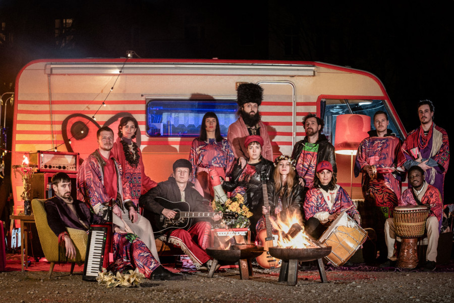 Das Bild zeigt den Caravan of LUV - insgesamt 12 Personen, männlich und weiblich gelesen vor einem Wohnwagen. Sie tragen bunte Kleider und haben verschiedene Musikinstrumente