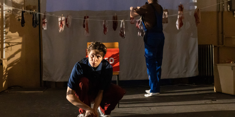 Das Bild zeigt zwei weiblich gelesene Personen in blauer Kleidung. Eine Person hockt im Vordergrund mit dem Gesicht nach vorn. Die zweite Person steht weiter hinten mit dem Rücken zur Kamera und hängt Binden an einer Leine auf.