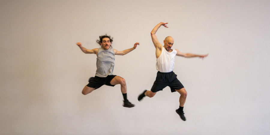 Das Bild zeigt zwei weiße, männlich gelesene Tänzer in der Luft. Bedie haben die Beine angewinkelt und die Arme über dem Kopf oder neben sich an den Seiten