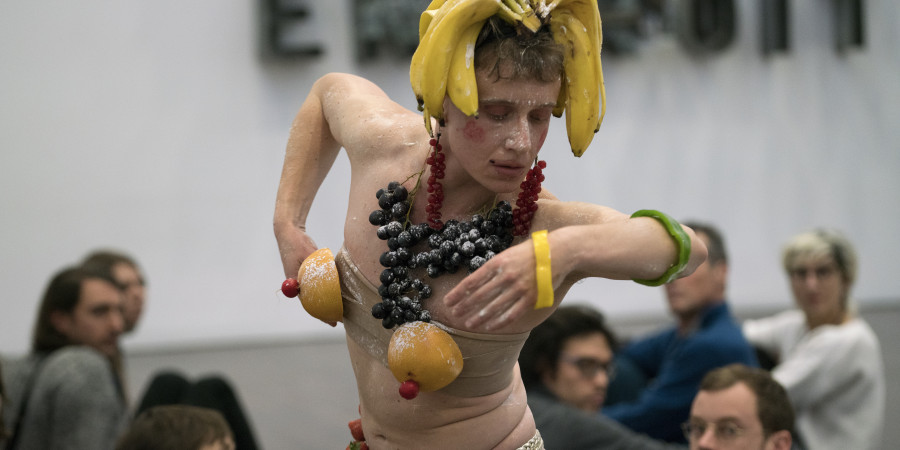 Das Bild zeigt den Performer Pol Pi, der mit Bananen auf dem Kopf, einem Collier aus Weintrauben und Brüsten aus Orangen geschmückt ist.