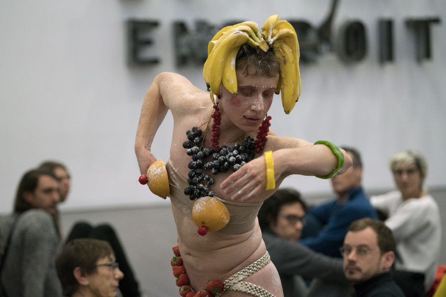 Das Bild zeigt den Performer Pol Pi, der mit Bananen auf dem Kopf, einem Collier aus Weintrauben und Brüsten aus Orangen geschmückt ist.
