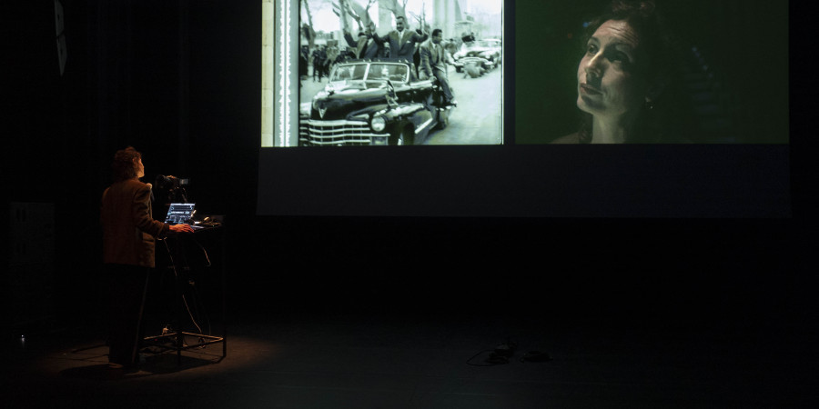 Das Bild ist ziemlich dunkel, im Hintergrund ist eine Leinwand zu sehen, auf der ein geteiltes Bild projeziert wird, links sind mehrere Männder in einem Auto zu sehen, daneben ist das Gesicht einer Frau.