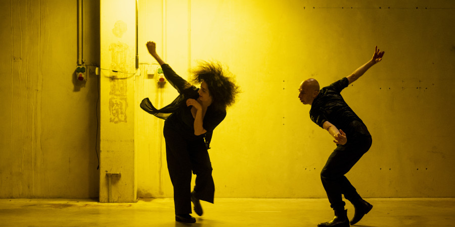 Das Bild zeigt die beiden Choreograf:innen Cristiana Morganti und Emanuele Soavi. Beide sind mitten in derBewegung, sie beugen sich vor, beide Arme gehen nach hinten. Das Licht in dem Foto ist sehr warm und wirkt gelb, der Ort wirkt industriell.