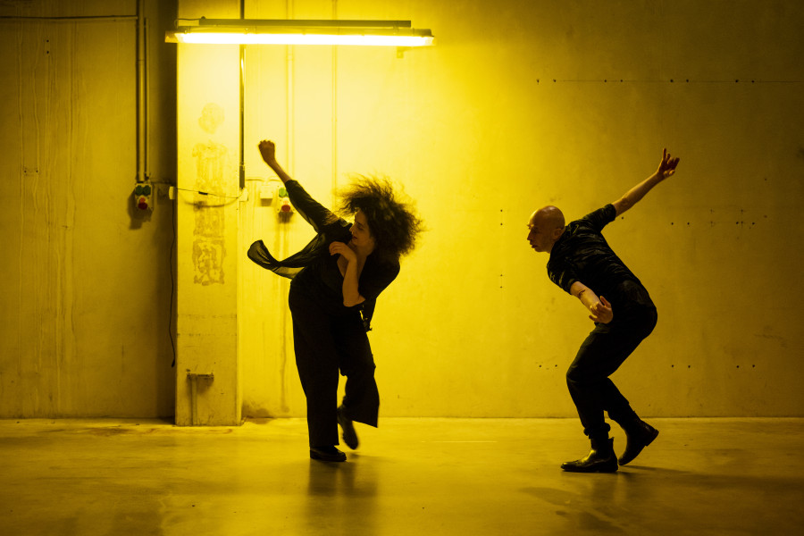Das Bild zeigt die beiden Choreograf:innen Cristiana Morganti und Emanuele Soavi. Beide sind mitten in derBewegung, sie beugen sich vor, beide Arme gehen nach hinten. Das Licht in dem Foto ist sehr warm und wirkt gelb, der Ort wirkt industriell.