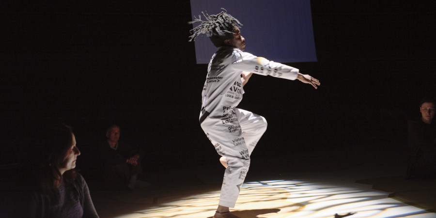 Das Bild zeigt den Tänzer Robert Ssempijja in weißer Kleidung. Er tanzt in einem erleuchteten Quadrat. An den Rändern kann man im dunkeln das Publikum erkannen, das auf allen Seiten Performance sitzt.