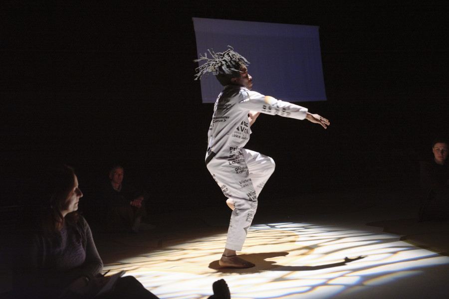 Das Bild zeigt den Tänzer Robert Ssempijja in weißer Kleidung. Er tanzt in einem erleuchteten Quadrat. An den Rändern kann man im dunkeln das Publikum erkannen, das auf allen Seiten Performance sitzt.